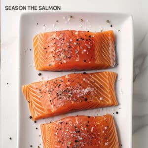 Image of a salmon fillet being generously seasoned on both sides with coarse salt and freshly cracked black pepper.