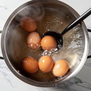 A slotted spoon adding a raw egg to pot of boiling water for boiling, preventing cracks.