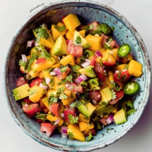 Close-up of fresh mango salsa featuring diced mango, red bell pepper, red onion, cilantro leaves, and lime wedges.