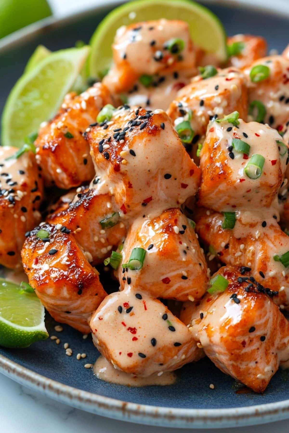 Crispy Bang Bang Salmon bites coated in a creamy, spicy sauce, garnished with sesame seeds, green onions, and red pepper flakes, served with lime wedges on a blue plate.